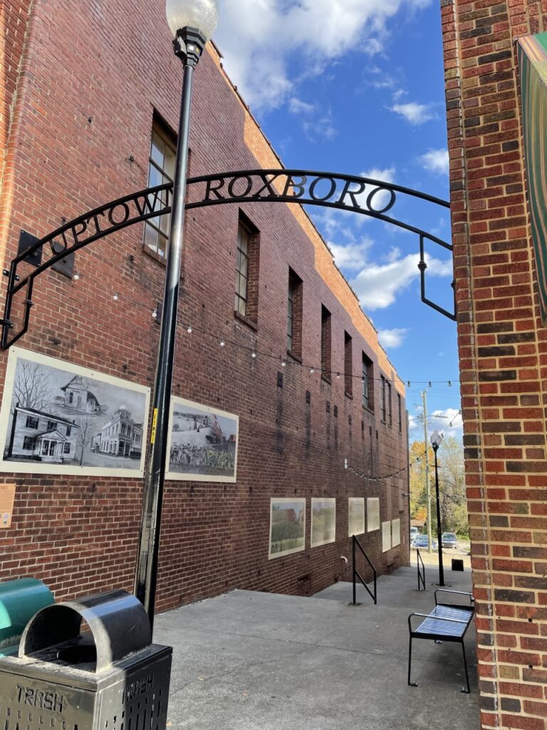 A black metal sign reading 'Uptown Roxboro' suspended between two red brick buildings